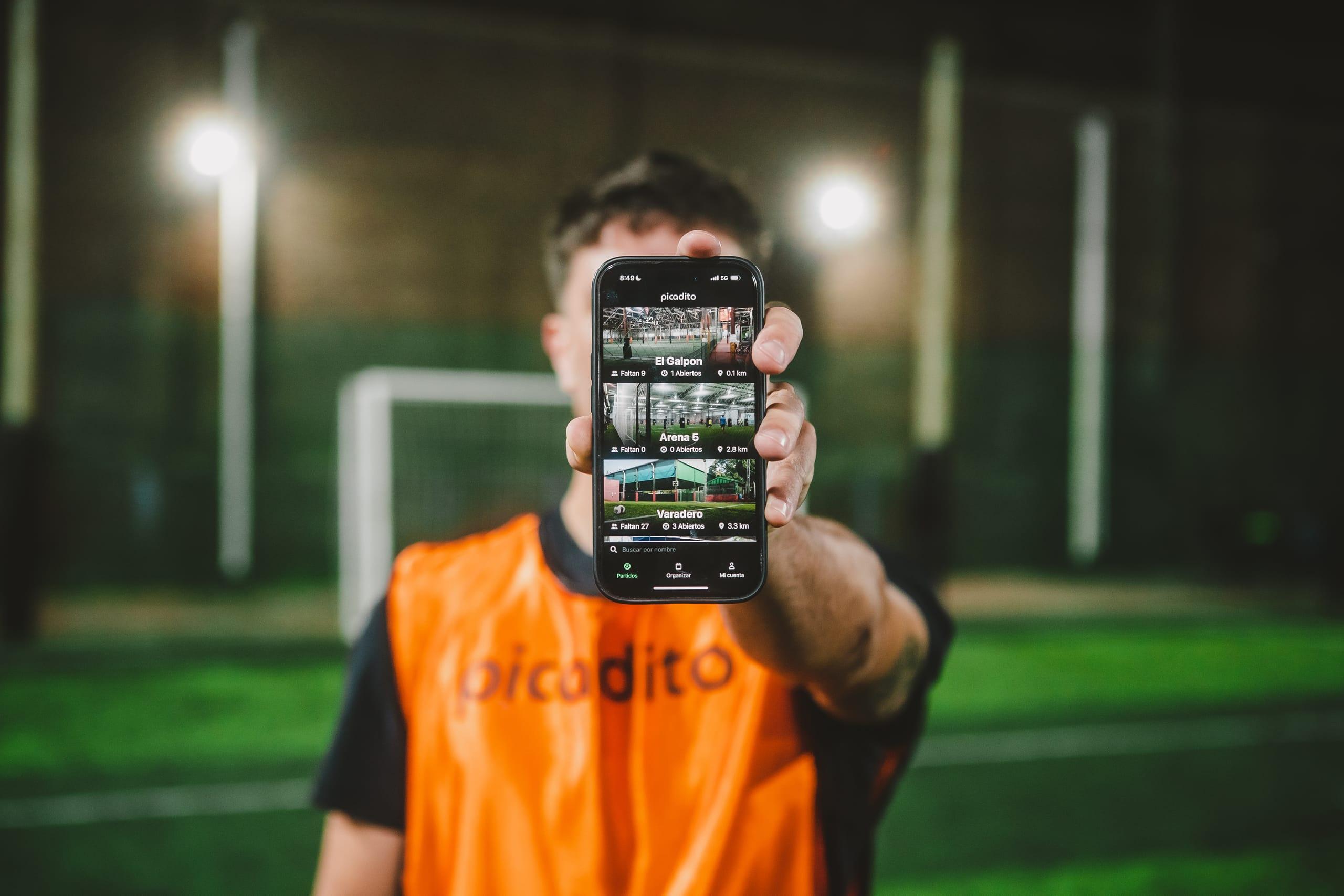 Celular con la lista real de complejos de picadito, con la cancha desenfocada detrás